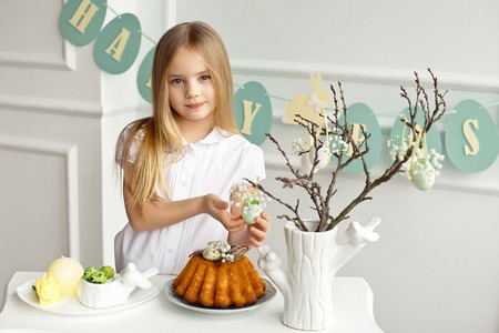 Beautiful blonde little girl in a white shirt is sitting at the holiday table near a vase with sprigs decorated with Easter eggs.の写真素材