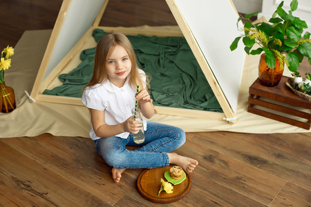 Cute happy little girl with blond hair in a white shirt and blue jeans is sitting near a tent with a glass bottle of lemonade and cookies with chocolateの写真素材