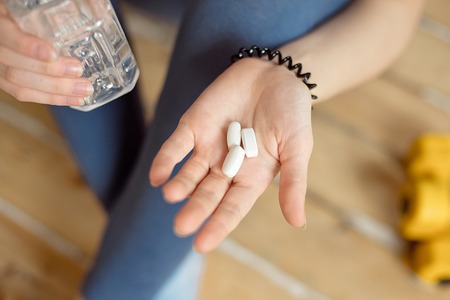 Sports girl in a blue shirt and blue leggings sits on the floor and holds in her hands sports supplements in capsules for bodybuilding.の写真素材