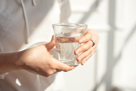 Close-up girl hands in the office stands in the sunlight holding a glass of clean water in between work.の写真素材