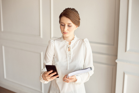 Photo business woman wearing suit, looking smartphone and holding documents in handsの写真素材