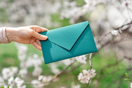 Close-up photo of female hands holding a green invitation envelope with a wax seal, a gift certificate, a postcard, a wedding invitation card on the background of blooming flowers.の写真素材