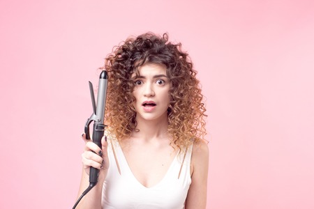 Beautiful woman with a surprised facial expression with amazing curls in a white shirt holds in her hand accessories for hair styling.の写真素材