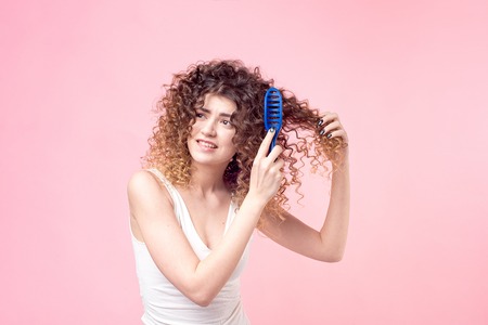 Beautiful woman with a smile on her face with curly hair in a white T-shirt combing blue curls with a blue comb isolate pink background.の写真素材