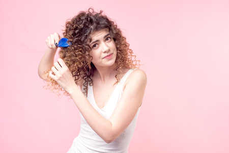 Beautiful woman with a smile on her face with curly hair in a white T-shirt combing blue curls with a blue comb isolate pink background.の写真素材