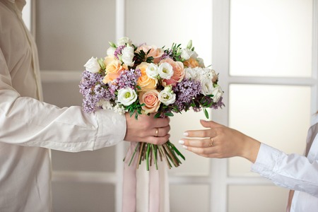 Man stretches out his hands and gives a large bouquet of flowers from roses and lilacs isolated against the background of a stained glass window. Close-up female hands take a bouquet. Gift for March 8th birthday weddingの写真素材