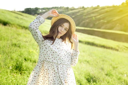 Beautiful sensual brunette in dress and hat standing with eyes closed in nature in bright back lit.の写真素材