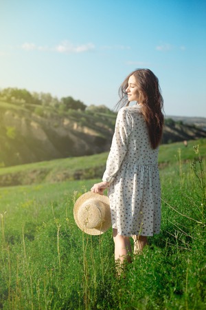 Beautiful sensual brunette in dress and hat standing with eyes closed in nature in bright back litの写真素材