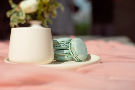 French macaroon blue plate on the pink and coffee cup standing on a wooden table with pink tablecloth white vase with flowers roses and greens.の写真素材