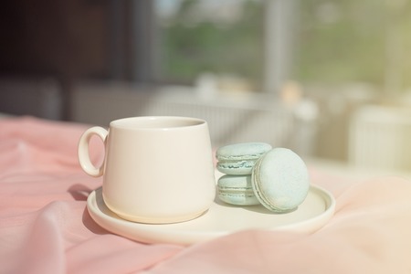 French macaroon blue plate on the pink and coffee cup standing on a wooden table with pink tablecloth white vase with flowers roses and greens.の写真素材