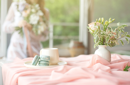 Young beautiful caucasian bride enjoying breakfast from french macaroon and coffee on a wooden table with a chiffon pink tablecloth and a vase of flowersの写真素材