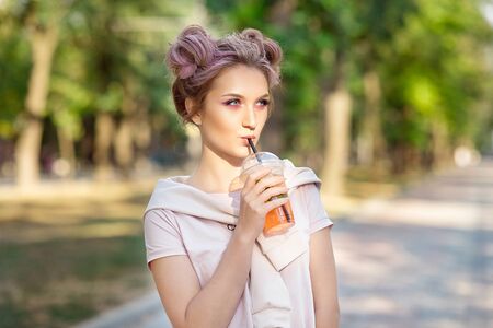 Young beautiful girl drinking fresh juice from plastic takeaway food cups after a walk outdoors. Healthy lifestyle. Smiling slim blonde with pink hair.の写真素材
