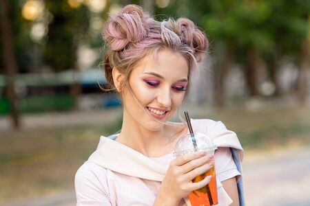 Young beautiful girl drinking fresh juice from plastic takeaway food cups after a walk outdoors. Healthy lifestyle. Smiling slim blonde with pink hairの写真素材