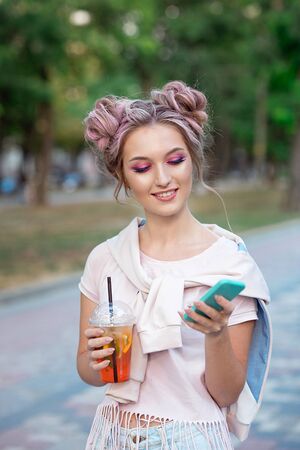 Young beautiful girl drinking fresh juice from plastic takeaway food cups after a walk outdoors. Healthy lifestyle. Smiling slim blonde with pink hairの写真素材