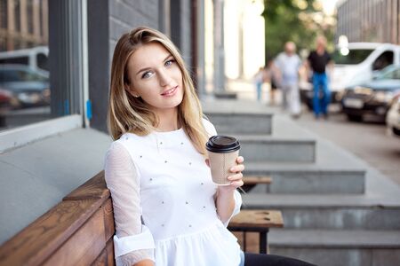 Beautiful girl walking in the city and drinking takes away coffee by an outdoor cafe. City morning scene.の写真素材