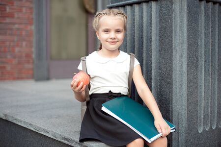 Happy little schoolgirl with blond hair in school uniform with lunch, book sits near schoolの写真素材