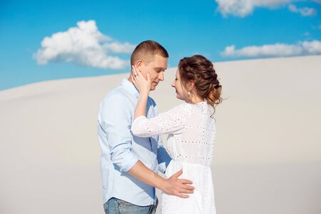 Photo of a gorgeous couple man and woman smiling and hugging on a sandy hill on a background of blue sky and white clouds.の写真素材