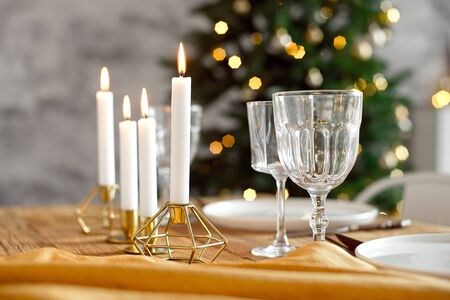Table is served for a Christmas dinner in the living room, close-up view, table setting with a golden tablecloth, white plates, forks, knives, glasses for wine, candles in gold candlesticks, Christmas decorationsの写真素材