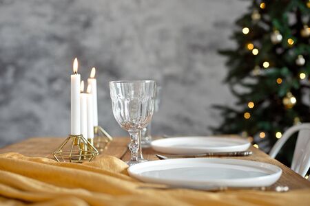 Table is served for a Christmas dinner in the living room, close-up view, table setting with a golden tablecloth, white plates, forks, knives, glasses for wine, candles in gold candlesticks, Christmas decorationsの写真素材