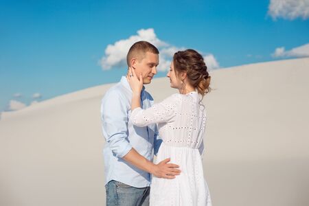 Photo of a gorgeous couple man and woman smiling and hugging on a sandy hill on a background of blue sky and white clouds.の写真素材
