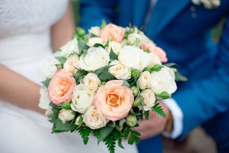 Close-up groom in a suit and the bride in a white dress are standing and holding a bouquet of peach roses, eustomas and flowers and greenery.の写真素材