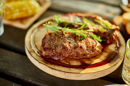 A succulent ribeye steak, perfectly grilled and adorned with fresh rosemary sprigs, rests on a circular wooden plate against a dark, rustic backdrop.の写真素材