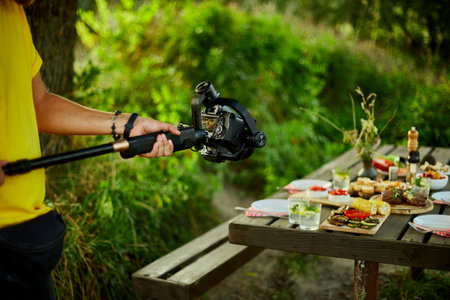 A person holds a camera on a stabilizing arm, filming a picnic table spread with food. The scene is outdoors, surrounded by greenery.の写真素材