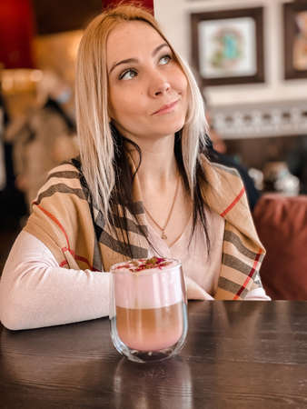 A blonde girl with a scarf on her shoulders in a soft pink sweater sits in a cozy restaurant and she is relaxed and have a coffee drink and a piece of cake on her table.の写真素材