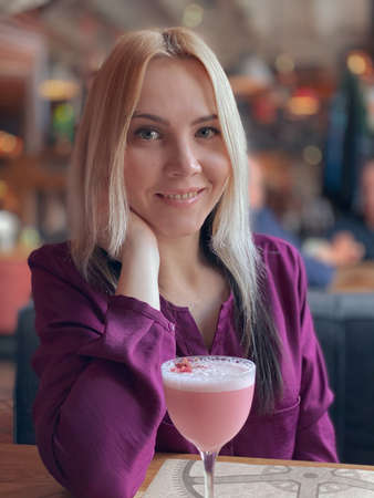 A blonde girl in a lilac sweater sits in a cozy restaurant on a sofa and she is relaxed and have a pink cocktail on her table.の写真素材