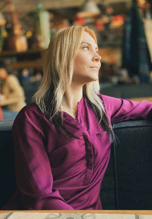 A blonde girl in a lilac sweater sits in a cozy restaurant on a sofa and she is relaxed and smiling.