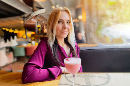 A blonde girl in a lilac sweater sits in a cozy restaurant on a sofa and she is relaxed and have a...
