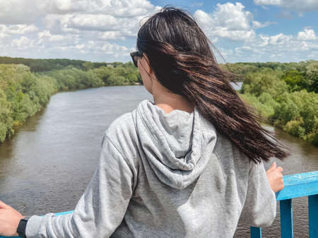 A dark haired caucasian girl stands with her back in a light gray jacket and looks towards the river and the forestの写真素材