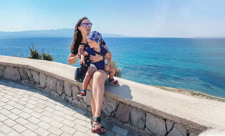 A dark haired caucasian woman with a baby in her arms on vacation is sitting against the backdrop of...