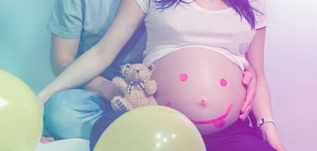Caucasian pregnant woman and a man who hugs her waist and balloons and a toy bear near themの写真素材