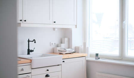 The interior of a kitchen in white tones with a sink, sandwich maker, stove, oven, dining table and...