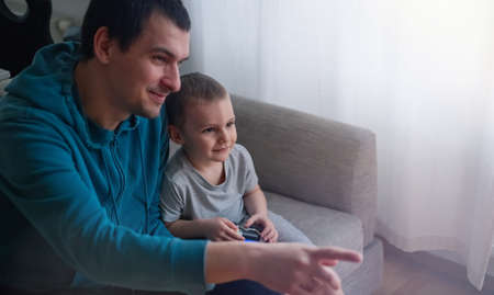 Happy father and son sit in the living room on the sofa and play or watch TV