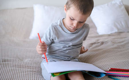 A small caucasian boy in a gray T-shirt and shorts on the bed draws with colored pencils in a sketchの写真素材