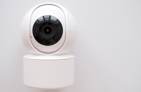 A white security camera with a black lens is mounted on a white surface. The camera is designed to...