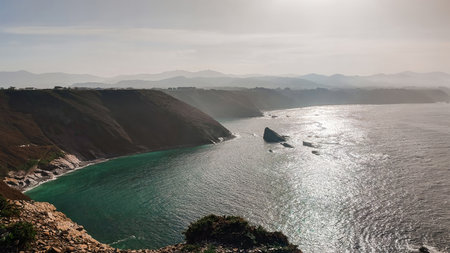 Coastal cliffs and shimmering sea under a soft hazy sky, with dramatic rock formations and distant mountains.の写真素材