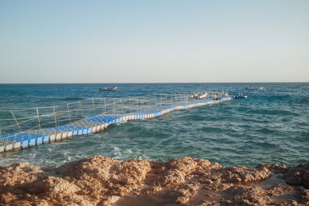 A tranquil view of a floating pier extending into the calm blue sea, with boats visible in the...