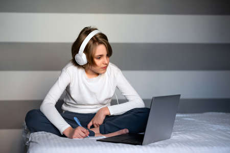 Teenager boy looking at laptop screen and writing at notebook, sitting on bed. Boy having lessons, studying online at home. distance education concept. home interior.の写真素材