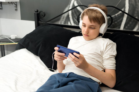 Teenager boy wearing earphones, laying on the bed with smartphone, playing games or watching videosの写真素材