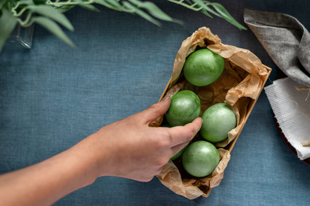 Green dyed Easter eggs in paper box on a blue background, hand taking an egg, spring holiday template with copy spaceの写真素材