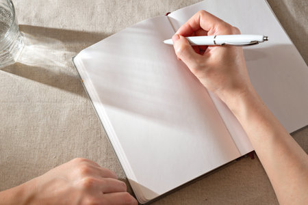 Women's hand writing in a blank notebook sheet with sunlight shadows, home office lifestyle workspace, mock up with copy spaceの写真素材