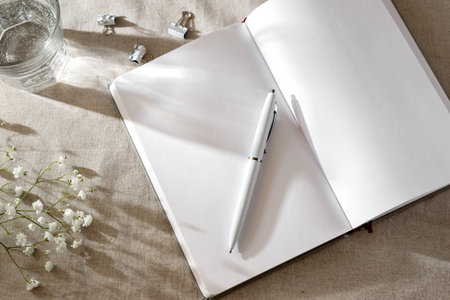 Open notebook blank sheets, pen and flowers on a neutral beige background with sunlight shadows,...