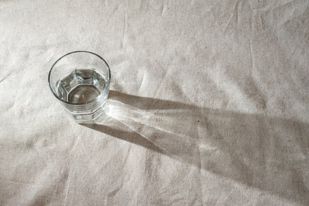 Glass of water on a beige linen tablecloth with sunlight shadows, minimalist aesthetic still life background with copy spaceの写真素材