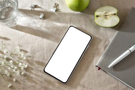 Mobile phone with empty white screen mock up, notebook, apples and flowers on a beige linen tablecloth, aesthetic spring or summer home workspaceの写真素材