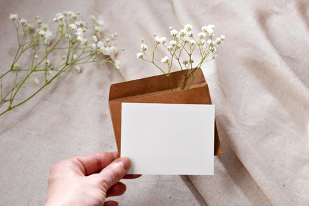 Blank paper card and envelope with flovers in persons hand on a beige background, elegant invitation, greeting card or business card template with mockup copy spaceの写真素材