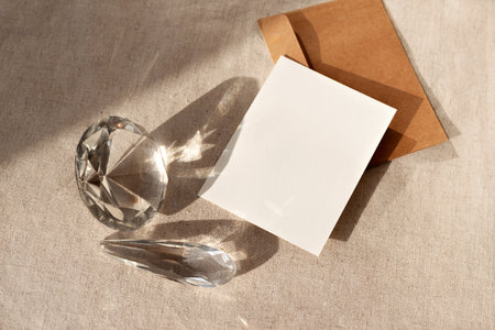 Blank paper sheet card with mock up copy space, crafted envelope and glass crystals with reflections and sunlight shadows on a neutral beige tablecloth, minimalist elegant business brand templateの写真素材