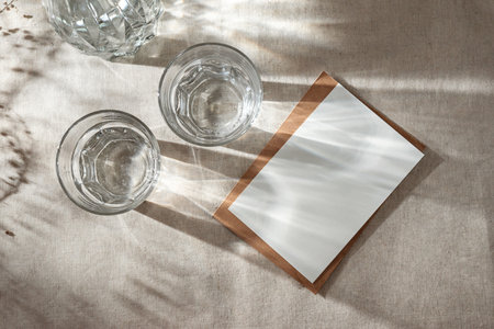 Blank paper card with mock up copy space, envelope, glasses of water and long harsh sunlight shadows on a neutral beige cloth, aesthetic business brand, invitation card templateの写真素材